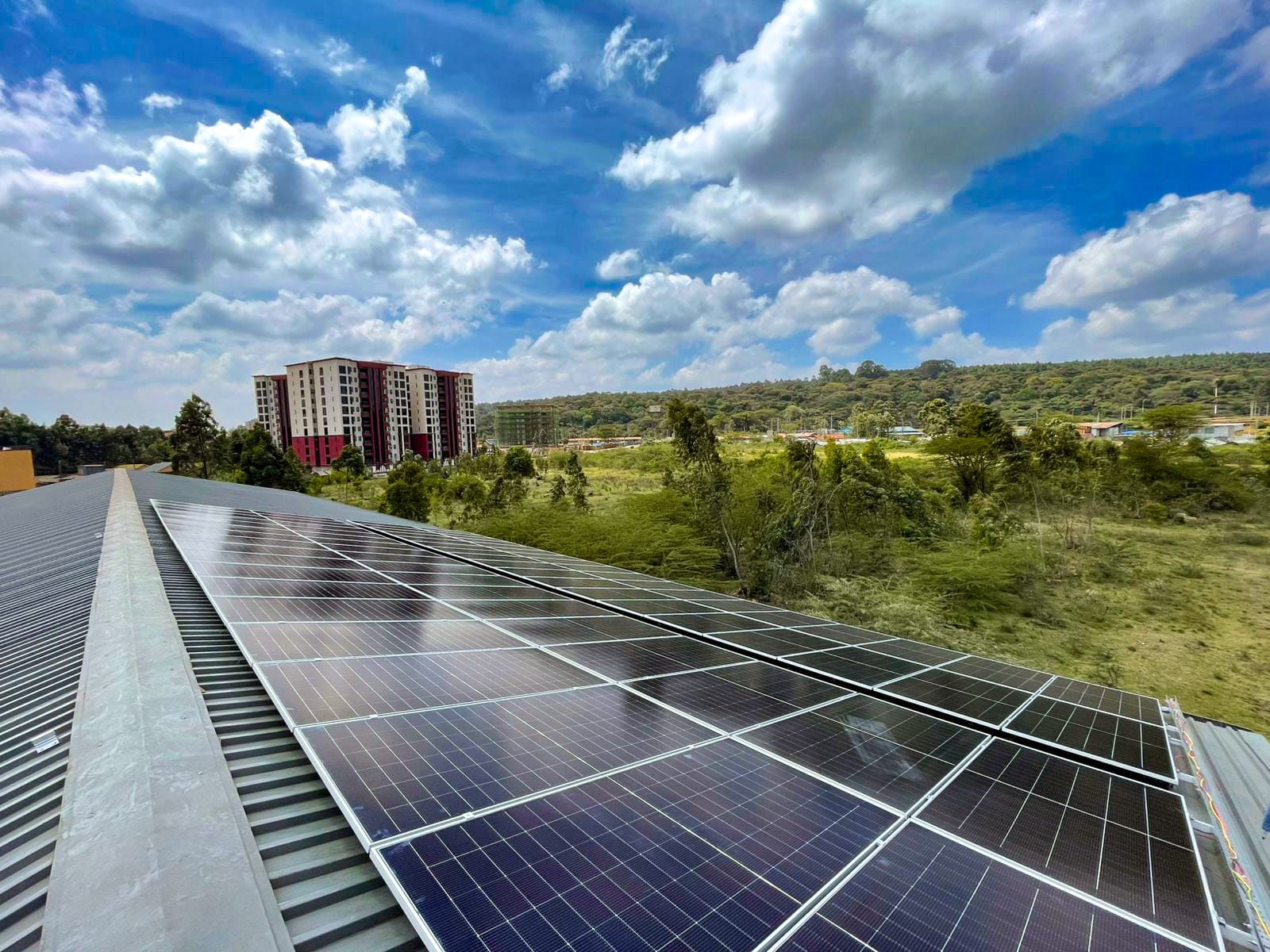 High-angle view of a large-scale solar panel installation on the roof of Almar Container Group's fabrication facility in Karen, Kenya, with modern apartment buildings and green landscapes in the background.